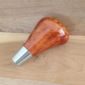 Könnte beinhalten: Ein Schaltknauf mit einem polierten, hochglänzenden, bernsteinfarbenen Holzmaserungsoberteil in einer glatten, abgerundeten, ergonomischen Form. Die Basis des Knaufs besteht aus einem konischen, gebürsteten, silberfarbenen Metallkragen.