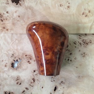 Könnte beinhalten: Ein glänzender Schaltknauf aus Holz mit einem satten, braunen Maserholz-Muster. Der Knauf hat eine glatte, abgerundete Oberseite und eine sich verjüngende Basis. Der Hintergrund ist eine hellfarbige Holzoberfläche mit einem einzigartigen, gesprenkelten Muster.