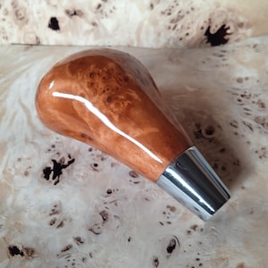 May include: A polished wooden gear shift knob featuring a rich, swirling burl grain pattern in shades of amber and brown, finished with a smooth, glossy surface and a tapered silver-toned metal base at the bottom for attachment.