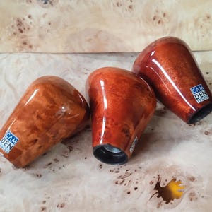 Pode incluir: Três manípulos de câmbio de madeira com acabamento brilhante. Os manípulos são de cor castanho-avermelhada com um padrão de grão de madeira. Cada manípulo tem uma base preta e uma pequena etiqueta azul e branca que diz "CAM DEN TUNING".