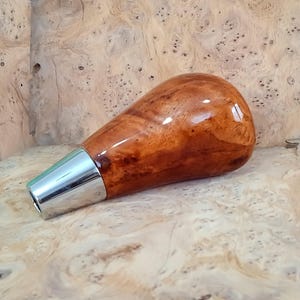 May include: A wooden gear shift knob with a brown and orange wood grain pattern and a silver metal base.