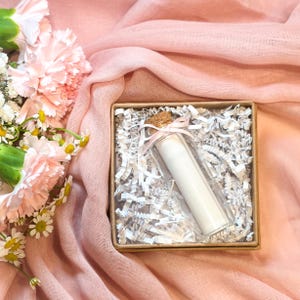 Könnte beinhalten: Eine klare Glasflasche mit Korken und rosa Schleife, die ein aufgerolltes weißes Papier enthält, befindet sich in einer kleinen Pappschachtel mit geschreddertem Papier. Die Schachtel liegt auf einem rosa Stoff, mit rosa und weißen Blumen im Hintergrund.