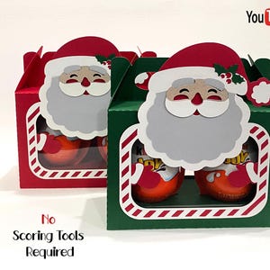 Może przedstawiać: Dwa świąteczne pudełka na prezenty, jedno czerwone i jedno zielone, każde z wycięciem w kształcie twarzy Świętego Mikołaja. Pudełka mają przezroczyste okienko pokazujące zawartość. Widoczny jest tekst "No Scoring Tools Required".