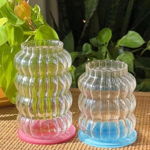 May include: Two clear glass vases with a wavy, textured design. The vases are sitting on colorful coasters. The larger vase is on a pink coaster and the smaller vase is on a blue coaster.