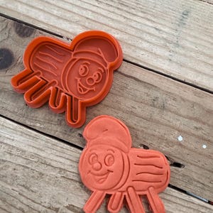 Puede incluir: Dos cortadores de galletas de plástico naranja con forma de personaje de dibujos animados con sombrero y patas. Los cortadores están sobre una superficie de madera.