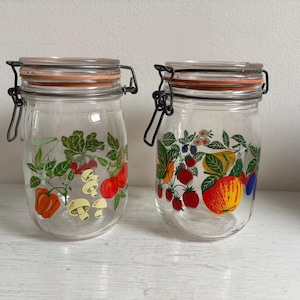 Op de afbeelding: Twee heldere glazen potten met scharnierende deksels. Eén pot heeft een groenteontwerp met tomaten, champignons en paprika's. De andere pot heeft een fruitontwerp met aardbeien, appels en bosbessen. De potten hebben een metalen sluiting.
