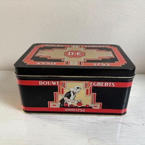 May include: Black rectangular tin with red and gold accents. The tin features the text "DOUWE EGBERTS" and "ANNO 1753". A central illustration depicts a person in a traditional Dutch outfit. The tin is likely for storing coffee or tea.