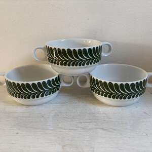Op de afbeelding: Drie witte keramische soepkommen met handvatten, elk versierd met een donkergroen bladerpatroon aan de buitenkant. De kommen zijn gestapeld en gerangschikt op een lichtgekleurd oppervlak.