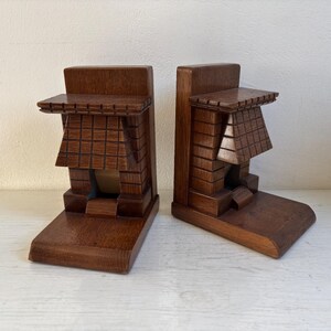 May include: Two wooden bookends in a dark brown hue, each with an architectural design. The bookends have a textured, geometric pattern, resembling a small building. The design is reminiscent of a miniature structure.