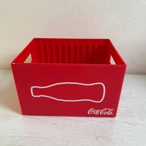 Könnte beinhalten: Eine rote Coca-Cola-Kiste aus Kunststoff mit einer weißen Umrisslinie einer Flasche auf der Vorderseite. Die Kiste hat eine rechteckige Form mit einem Griff auf jeder Seite und dem Coca-Cola-Logo in Weiß.