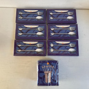 Puede incluir: Seis cajas de cucharas, tenedores y cucharas decorativas plateadas con varios diseños, incluyendo peces, corazones y conchas. Las cajas son de color azul oscuro con texto blanco. También se ve un paquete separado de cucharas.
