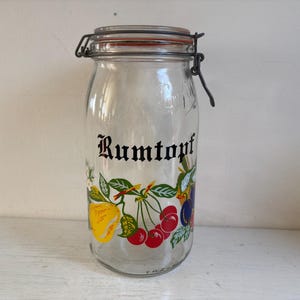 May include: Clear glass jar with a metal hinged lid and a red rubber seal. The jar is decorated with a colourful fruit design and the word "Rumtopf" in black script.