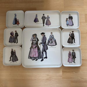 Op de afbeelding: Een verzameling witte keramische trays en borden met illustraties van mensen in historische kleding. De afbeeldingen tonen stellen en gezinnen in verschillende poses, met gedetailleerde kleding en accessoires. De trays zijn rechthoekig en vierkant.