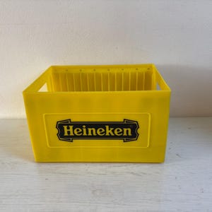 Könnte beinhalten: Eine gelbe Plastikkiste mit dem Heineken-Logo in Schwarz und Gold. Die Kiste hat eine rechteckige Form mit einer Reihe von vertikalen Trennwänden im Inneren. Sie hat zwei Grifföffnungen an den Seiten.