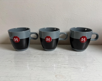 Op de afbeelding: Drie grijze en zwarte koffiemokken met een rood embleem met de letters "D-E". De mokken hebben een afgeronde vorm met een oor en zijn in een rij op een wit oppervlak geplaatst. De mokken hebben een tweekleurig ontwerp.