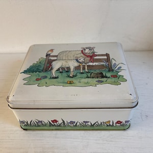 Op de afbeelding: Een vintage witte metalen doos met een deksel met een pastorale scène. Het deksel toont twee schapen, een hek en een vogel. De basis van de doos is versierd met een groene rand van gras, bloemen en vogels.