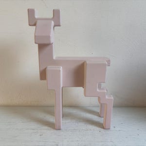 Op de afbeelding: Een lichtroze, geometrisch hertenbeeld. Het hert is opgebouwd uit rechthoekige en vierkante blokken, waardoor het een gepixeleerd uiterlijk heeft. De sculptuur heeft een eenvoudig, minimalistisch ontwerp en staat op vier poten. De geweien zijn ook rechthoekig.