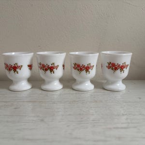 Op de afbeelding: Vier witte eierdopjes met een bloemmotief. Elke beker heeft een voet en een uitlopende bovenkant. Het bloemmotief toont rode en roze rozen met groene bladeren. De bekers zijn in een rij gerangschikt.