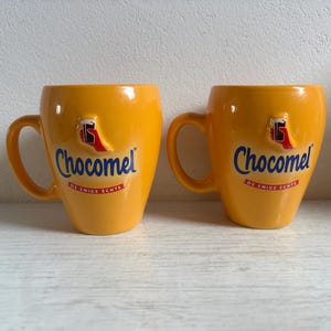 Könnte beinhalten: Zwei leuchtend gelbe Keramiktassen mit dem blauen Chocomel-Logo und einer kleinen erhabenen Grafik einer Hand, die ein Glas hält. Unter dem Logo steht der Text DE ENIGE ECHTE in Rot und Weiß. Jede Tasse hat einen geschwungenen Henkel und Oberfläche.