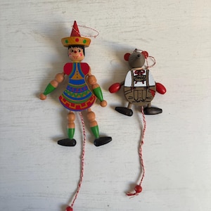 May include: Two wooden pull-string ornaments. One is a figure wearing a red hat, blue dress with colorful patterns, and green sleeves. The other is a brown mouse wearing white and brown lederhosen with the name Freddy written on the chest.