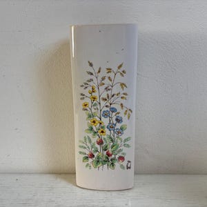 Peut inclure: Un vase en céramique blanc cassé, haut et au profil plat et rectangulaire. La surface avant présente une illustration botanique détaillée de fleurs sauvages, d'herbes et de petites baies rouges dans des tons de jaune, bleu, vert et rouge.