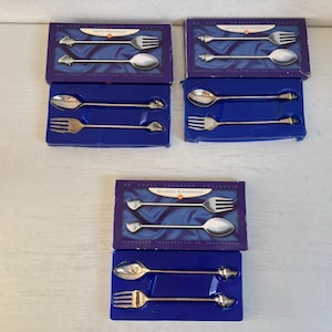 Op de afbeelding: Besteksets met lepels en vorken, elk met een decoratief uiteinde. De sets zijn verpakt in blauwe dozen met paarse deksels. De dozen hebben Duitse tekst. Het bestek heeft een nautisch thema.