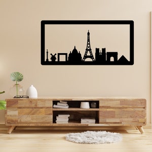 Op de afbeelding: Zwart-witte silhouet van de skyline van Parijs, inclusief de Eiffeltoren, de Arc de Triomphe en de Notre Dame kathedraal, weergegeven in een zwart frame op een beige muur boven een houten entertainmentcenter.
