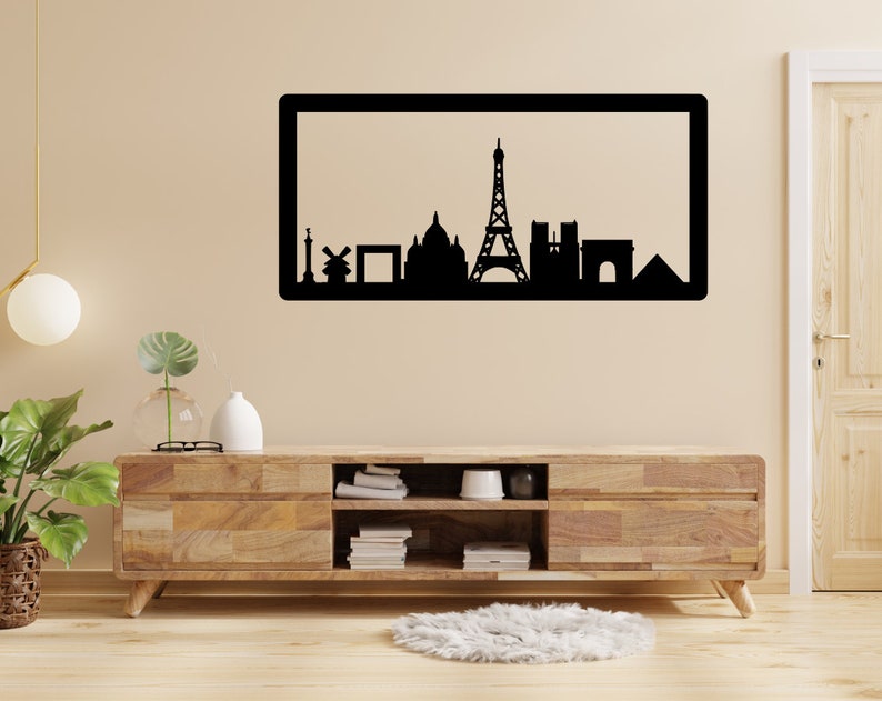 Op de afbeelding: Zwart-witte silhouet van de skyline van Parijs, inclusief de Eiffeltoren, de Arc de Triomphe en de Notre Dame kathedraal, weergegeven in een zwart frame op een beige muur boven een houten entertainmentcenter.