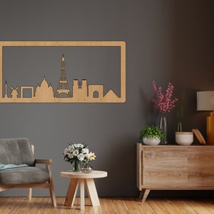 Op de afbeelding: Een houten wanddecoratie met een silhouet van de skyline van Parijs, inclusief de Eiffeltoren, de Arc de Triomphe en andere iconische bezienswaardigheden. De decoratie is gemonteerd op een grijze muur boven een houten dressoir met een lamp en bloemen.