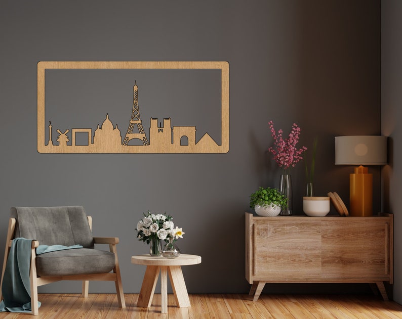 Op de afbeelding: Een houten wanddecoratie met een silhouet van de skyline van Parijs, inclusief de Eiffeltoren, de Arc de Triomphe en andere iconische bezienswaardigheden. De decoratie is gemonteerd op een grijze muur boven een houten dressoir met een lamp en bloemen.