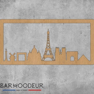 Op de afbeelding: Een houten uitsnijding van de skyline van Parijs, met iconische bezienswaardigheden zoals de Eiffeltoren, de Arc de Triomphe en de Notre Dame kathedraal. De uitsnijding is gemonteerd op een lichtbruin houten frame.