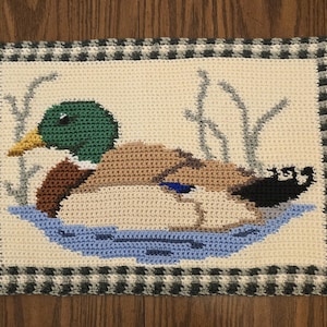 May include: Crocheted rectangular wall hanging featuring a mallard duck in a pond. The duck has a green head, brown body, and blue accents. The background is cream-colored with gray reeds, and a black and white checkered border.
