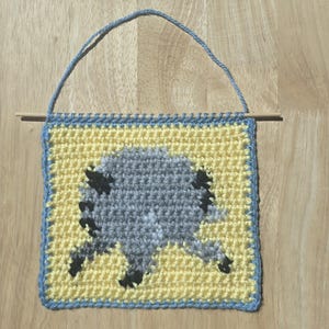 May include: A square crochet wall hanging featuring a yellow background with a central gray and black sheep graphic. The piece is bordered in light blue, suspended from a thin wooden dowel with a matching light blue yarn hanging loop.