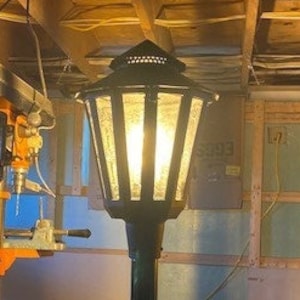May include: A black street lamp with a glass shade and a metal frame is mounted on a black pipe base. The lamp is sitting on a wooden table with a drill press and a saw in the background.