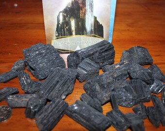 Può includere: Una collezione di cristalli di turmalina nera, alcuni con striature, su una superficie di legno. I cristalli sono di varie dimensioni e forme, alcuni sono lunghi e sottili, mentre altri sono più arrotondati. I cristalli sono di colore nero scuro, e alcuni hanno una leggera lucentezza. Il testo "BLACK TOURMALINE" è visibile su una carta sullo sfondo.