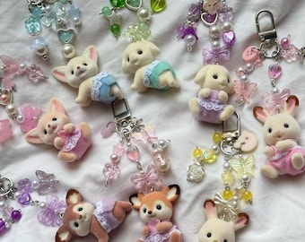 May include: A collection of keychains featuring flocked animal figures in pastel-colored clothing, each attached to a metal clasp and decorated with various beads, including translucent butterflies, hearts, stars, and round pearls in diverse colors.
