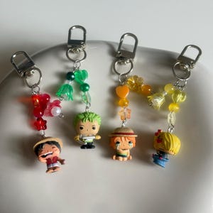 May include: Four keychains featuring miniature anime character figures attached to silver-tone metal lobster clasps. Each keychain is decorated with colourful beads in red, green, orange, and yellow, including heart, star, and flower-shaped accents.