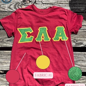 Könnte beinhalten: Ein rotes T-Shirt mit grünen und gelben griechischen Buchstaben. Die Buchstaben sind mit einem Paisley-Muster bestickt. Das Shirt hat den Text "CUSTOM GREEK LIFE LETTERS!" in einem rosa Banner. Lizenziertes Produkt.