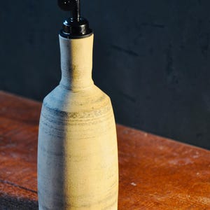 May include: A ceramic olive oil bottle with a black spout. The bottle is a light beige colour with horizontal grey stripes. The bottle is on a wooden surface.