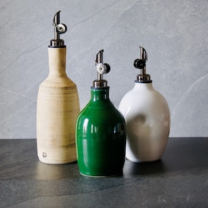 Könnte beinhalten: Drei Keramikflaschen für Öl und Essig mit Ausgießern aus Edelstahl. Die Flaschen sind in Creme, Grün und Weiß gehalten. Die cremefarbene Flasche hat eine strukturierte Oberfläche. Die grüne Flasche ist glänzend.