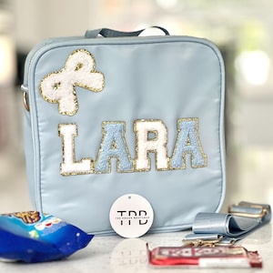 May include: A light blue lunch bag with the name "LARA" in white and light blue letters. The bag has a gold bow and a matching strap. A tag reads "THE POUCH BOUTIQUE". A snack and candy bar are visible.