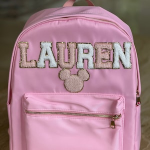 May include: Pink backpack with a gold zipper and a white, pink and gold "LAUREN" name patch with a pink and gold Mickey Mouse patch. The backpack has a small zipped pocket on the front.