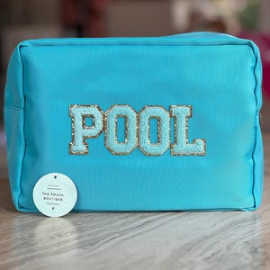 Może przedstawiać: Turkusowa prostokątna kosmetyczka z napisem "POOL" w jasnoniebieskich, cekinowych literach. Kosmetyczka ma małą, okrągłą, białą metkę z czarnym napisem "THE POUCH BOUTIQUE". Kosmetyczka leży na jasnobrązowej drewnianej powierzchni.
