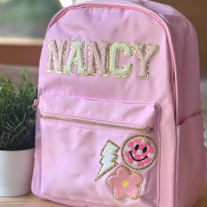 May include: Pale pink backpack featuring the name "NANCY" in gold and cream-coloured letters. The bag has a front pocket with a gold zip and decorative patches, including a lightning bolt, a smiley face, and a flower.
