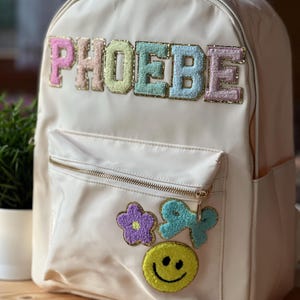 Può includere: Zaino color crema con il nome "PHOEBE" in lettere strutturate dai colori pastello. Lo zaino presenta una tasca frontale con cerniera dorata e toppe decorative, tra cui un fiore, un fiocco e una faccina sorridente.