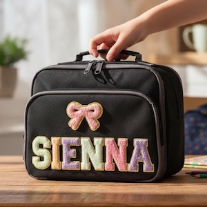 May include: Black lunch bag with a bow and the name "SIENNA" in colourful letters. The bag has a handle and a zip closure. A hand is holding the bag on a wooden table.