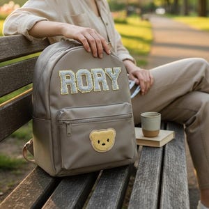 Może przedstawiać: Taupe plecak z napisem "RORY" dużymi, błyszczącymi literami. Plecak posiada przednią kieszeń z naszywką z misiem. Mały kubek i książka leżą obok plecaka. Osoba ma na sobie beżowy strój.