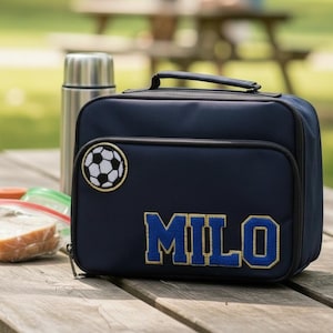 May include: Navy blue lunch bag with a soccer ball patch and the name "MILO" in blue and gold lettering. A stainless steel thermos, apple, sandwich, and other snacks are on a wooden picnic table.