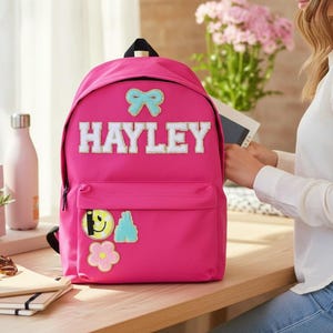 Può includere: Uno zaino rosa acceso con il nome "HAYLEY" in lettere bianche. Presenta un fiocco azzurro, una faccina sorridente, un fiore e una toppa a forma di montagna. Una tasca frontale e una maniglia nera completano il design.