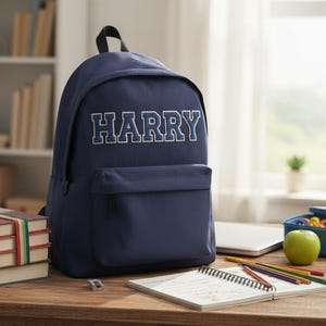 May include: A navy blue backpack with the name "HARRY" in white block letters. The backpack has a front pocket and a top handle. A stack of books, pencils, and an apple are on the wooden desk.